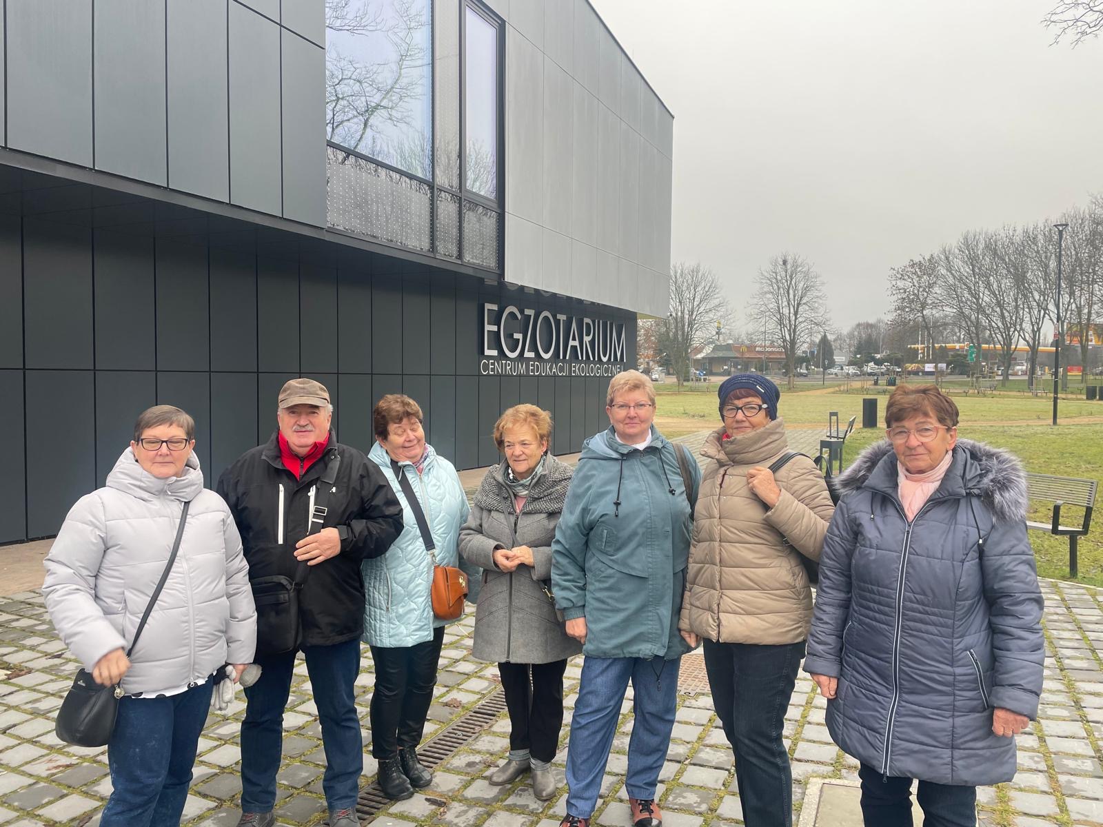 Grupa seniorów stoi w rzędzie i pozuje do zdjęcia, w tle budynek Egzotarium w Sosnowcu.jpeg